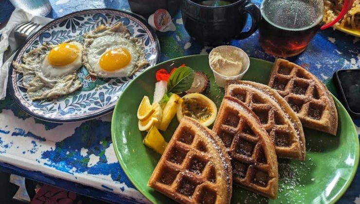 A plate of two sunny-side-up eggs sits on the left, next to a plate of four waffle quarters dusted with powdered sugar on the right. The waffles are accompanied by lemon slices, serving of butter, and a garnish. A pitcher of syrup and a mug are in the background.