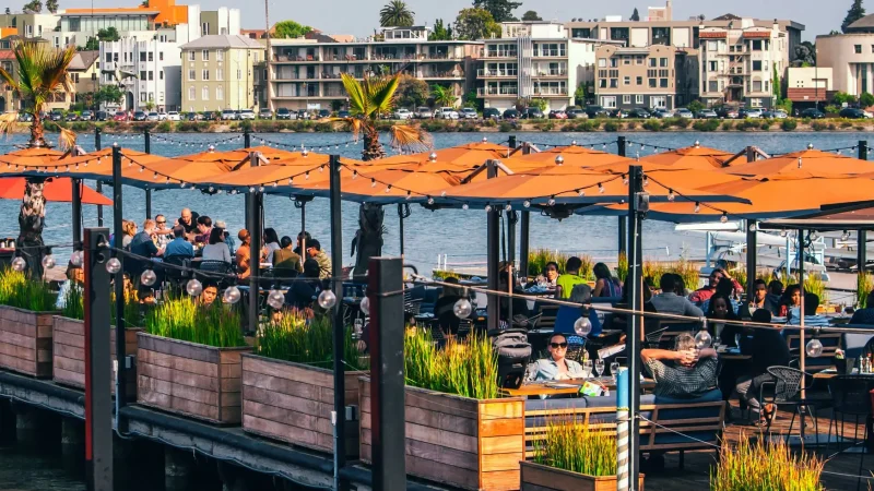 A bustling outdoor waterfront restaurant with orange umbrellas, wooden planters, and string lights. Patrons enjoy food and drinks while overlooking a body of water. Across the water, various buildings and lush green trees are visible under a clear sky.