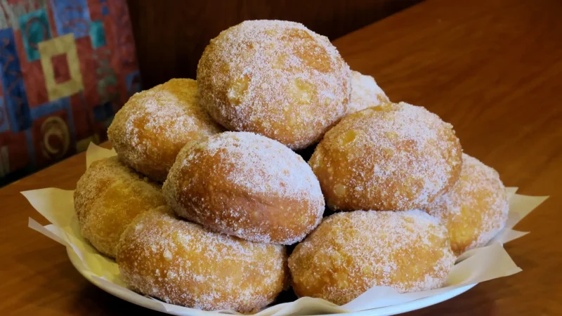 A plate filled with several sugar-coated doughnuts is placed on a wooden table. The doughnuts are stacked into a small mound, creating an appealing display of freshly baked treats.