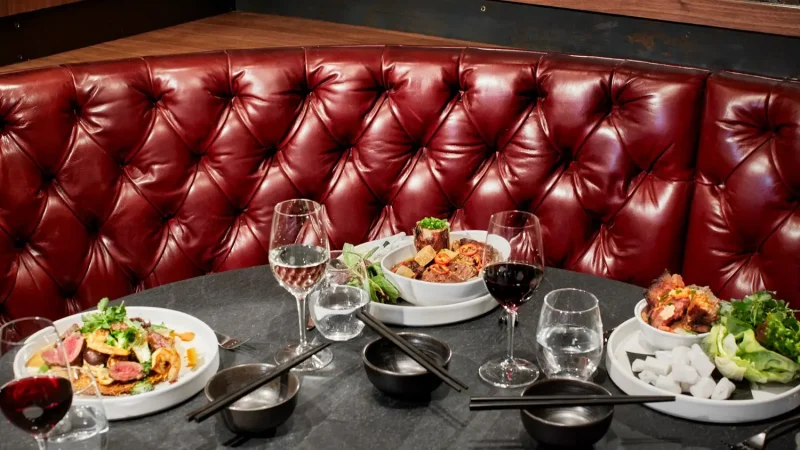 A restaurant booth with a luxurious red-tufted leather banquette. On the table are elegant dishes with diverse food items, including pasta and salads, along with several wine glasses filled with white and red wine, and bowls with chopsticks.