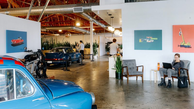 A coffee shop building with tables, people sitting and walking around, art on the walls and old cars inside on display.