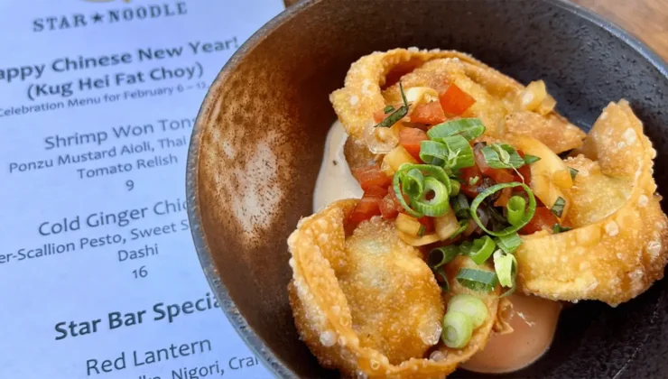 star-noodle-kaanapali-lunch-under-20-@starnoodle-facebook-1600×800-1.png A bowl of crispy shrimp won tons garnished with chopped green onions and diced tomatoes is placed on a table. A menu featuring various dishes, including the shrimp won tons, is partially visible in the background. The event is for a Chinese New Year celebration.